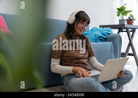 Glückliche lateinfrau, die Kopfhörer trägt und einen Laptop benutzt, während sie zu Hause auf dem Boden sitzt Stockfoto
