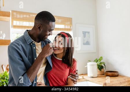 Ein vielseitiges Paar, das einen verspielten Moment zu Hause genießt, lächelt und in der Küche umgeht Stockfoto