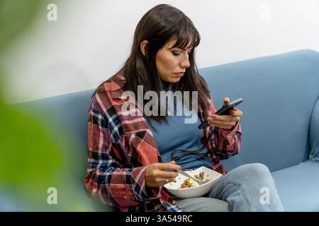 Junge Frau, die ihr Smartphone benutzt und Salat isst, während sie zu Hause auf dem Sofa sitzt Stockfoto
