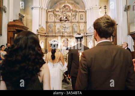 Montenegro, Kotor - 13. Juli 2024: Gäste stehen in der Kirche hinter Braut und Bräutigam in Kränzen vor dem Priester während der Hochzeitszeremonie Stockfoto