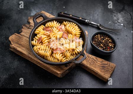 Fusilli Carbonara Nudeln mit knusprigen Speckstückchen und Parmesankäse in einer Pfanne. Schwarzer Hintergrund. Draufsicht. Stockfoto