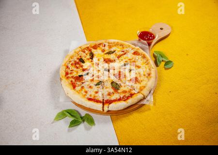 Pizza Margarita, gebacken in einem Holzofen, serviert auf einem Holzteller mit Basilikumblättern und Pizzasauce auf der Seite auf weißem und gelbem Hintergrund. Stockfoto