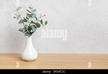 Eukalyptuszweige, die in einer weißen Vase auf einem Holztisch vor einer strukturierten Wand angeordnet sind, schaffen eine frische und natürliche Atmosphäre Stockfoto