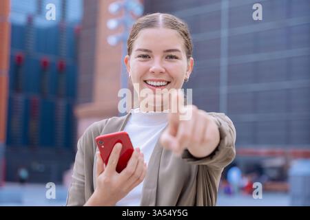 Lächelnde weiße junge Frau, die draußen mit rotem Smartphone auf die Kamera zeigt Stockfoto