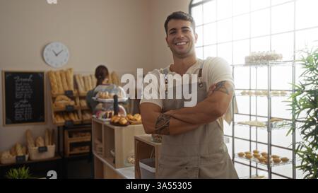 Ein Mann mit Tätowierung steht selbstbewusst mit überkreuzten Armen in der Bäckerei und zeigt Brot und Gebäck, während die Frau im Hintergrund arbeitet und die es festnimmt Stockfoto