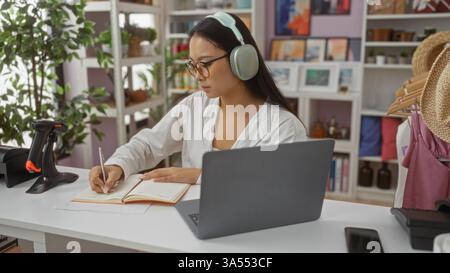 Frau, die in einem Notizbuch auf dem Schreibtisch im Dekorationsgeschäft, umgeben von Pflanzen und modernen Dekorationselementen, mit Kopfhörern und Brillen, die sich auf Aufgaben konzentrieren, schreibt Stockfoto