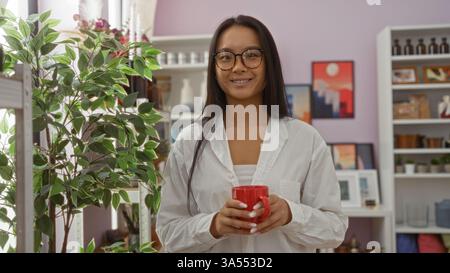 Junge chinesen, eine Frau, die einen roten Becher hält, steht in einem Haus-Dekorationsgeschäft, umgeben von Innenpflanzen und verschiedenen Dekorationen in Regalen. Stockfoto