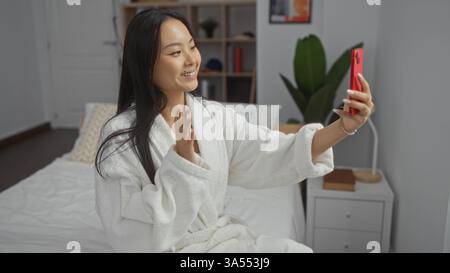 Frau Videoanrufe aus dem gemütlichen Schlafzimmer, trägt Bademantel, lächelt, während sie mit dem modernen Smartphone in der Hand auf dem Bett sitzt und die entspannte Umgebung in ihrem Zuhause festnimmt Stockfoto