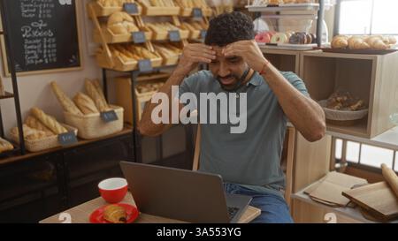 Junger Mann, der drinnen in einem Bäckereicafé mit einem Laptop und Kaffee sitzt, umgeben von Brot und Gebäck, das gestresst aussieht Stockfoto