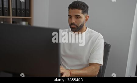 Hübscher junger Mann, der an einem Computer in einem Büro arbeitet, sich mit einem neutralen Ausdruck auf seine Aufgaben konzentriert, vor dem Hintergrund des organisierten Schwarzen Stockfoto
