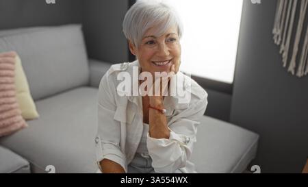Schöne grauhaarige Frau mit kurzen Haaren lächelt in einem Wohnzimmer, zeigt einen ruhigen und fröhlichen Ausdruck, drinnen in einer gemütlichen häuslichen Umgebung mit Stockfoto