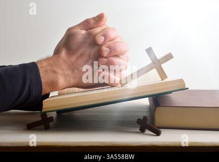 Katholischer Mensch betet mit Händen über einer Bibel auf einem Holztisch mit hölzernen religiösen Kreuzen und weißem isolierten Hintergrund mit Witz Stockfoto