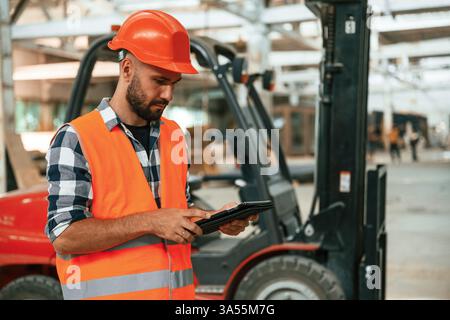Der Gabelstapler ist hinter dem Gerät, das digitale Tablet in den Händen. Industriearbeiter im Holzlager. Stockfoto