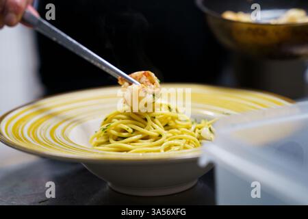 Ein Küchenchef legt gegrillte Garnelen vorsichtig mit einer Metallzange auf einen Teller Spaghetti aglio e olio. Die Pasta, die mit Olivenöl glänzt, wird garniert Stockfoto