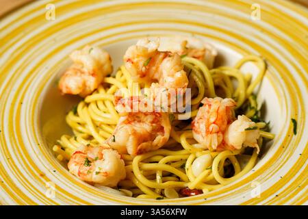 Ein wunderschön überzogener Garnelen-Spaghetti-aglio e olio wird auf einer gelb gestreiften Keramikschale auf einem Holztisch präsentiert. Die Teigwaren sind mit Olivenöl überzogen Stockfoto