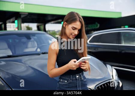 Mit dem Smartphone durch das Internet scrollen. Eine junge Frau an einer Tankstelle mit ihrem Auto. Stockfoto