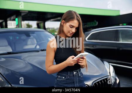 Mit dem Smartphone durch das Internet scrollen. Eine junge Frau an einer Tankstelle mit ihrem Auto. Stockfoto