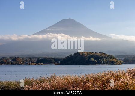 Der Fuji erhebt sich über dem See Kawaguchi in Japan, teilweise bedeckt von Wolken während der goldenen Stunde, Herbstlaub färbt die Bäume auf der Insel Stockfoto