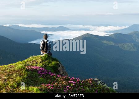 Atemberaubende Berglandschaft mit einem Reisenden, der zwischen blühenden Rhododendronen sitzt und den Sonnenaufgang über nebelnden Karpaten beobachtet. Frühlingsberge. Landschaftsfotografie Stockfoto