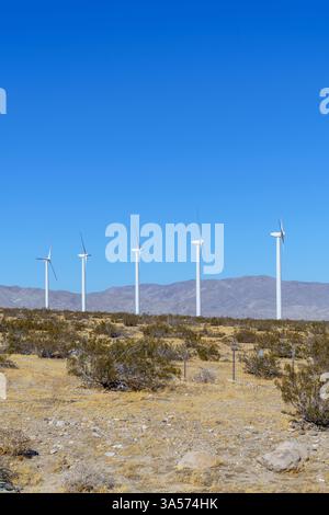 Fünf weiße Windräder stehen in einer Reihe vor einem klaren blauen Himmel und einer trockenen Wüstenlandschaft von Palm Springs. Im Hintergrund sichtbare Berge mit c Stockfoto