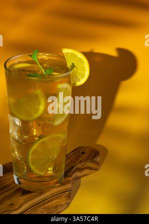 Ein Glas Eistee mit Limetten- und Zitronenscheiben darin. Das Getränk wird auf dem Holzbrett in einem Glas mit Minzblättern auf der Oberseite serviert. Stockfoto