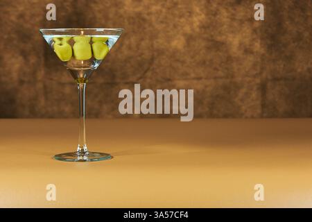 Erfrischendes Martini-Glas mit grünen Oliven im Inneren, auf einer warmen farbigen Oberfläche. Stockfoto