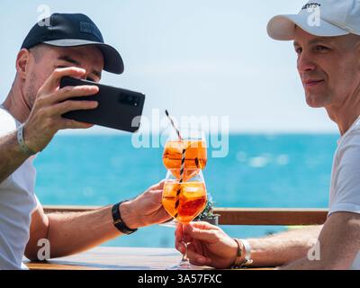 Fuengirola, Malaga, Spanien .03/15/2025. Schwule Paare machen ein Selfie mit ihrem Handy und toasten mit einem Cocktail vor dem Meer. Stockfoto