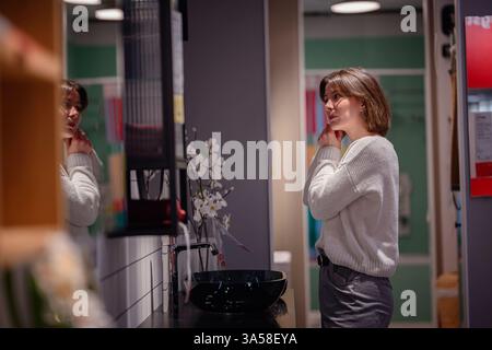 Eine Frau in einem weißen Pullover untersucht ihre Reflexion in einem Spiegel, während sie einen Ausstellungsraum im Badezimmer in einem Wohnhaus erkundet. Stockfoto