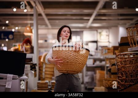 Eine Frau in einem weißen Pullover untersucht einen großen gewebten Korb in einem Heim-Geschäft und zieht ihn zum Aufbewahren oder Dekorieren beim Einkaufen von Haushaltsgegenständen in Betracht. Stockfoto