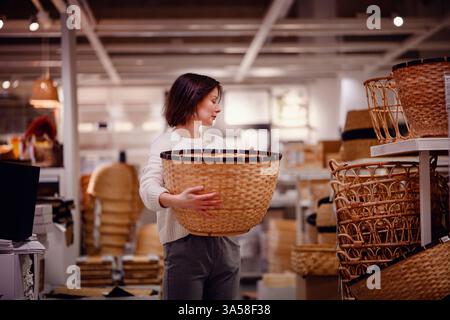 Eine Frau in einem weißen Pullover untersucht einen großen gewebten Korb in einem Heim-Geschäft und zieht ihn zum Aufbewahren oder Dekorieren beim Einkaufen von Haushaltsgegenständen in Betracht. Stockfoto