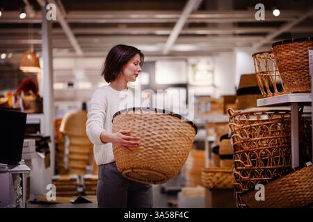 Eine Frau in einem weißen Pullover untersucht einen großen gewebten Korb in einem Heim-Geschäft und zieht ihn zum Aufbewahren oder Dekorieren beim Einkaufen von Haushaltsgegenständen in Betracht. Stockfoto