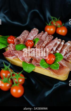 Flache Lagen verschiedener italienischer Wurstwaren, einschließlich Schinken, Salami und coppa, kunstvoll auf einem Holzbrett mit frischen Basilikumblättern und Kirschtomaten angeordnet. Der tiefschwarze satinierte Hintergrund verstärkt die satten Farben und Texturen der Zutaten und schafft eine raffinierte und appetitliche Präsentation. Konzept für Gourmetküche, Catering, Restaurantmenü und Lebensmittelfotografie. Stockfoto
