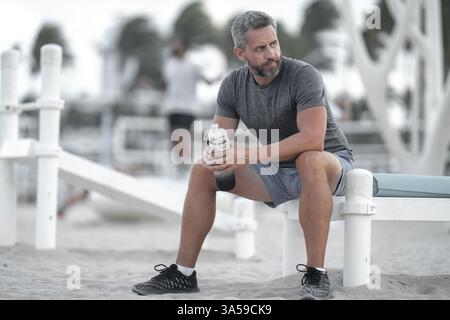 Sport-Fitness-Mann, der draußen auf einer Bank ruht. Reifes Fitnesssportmodell, das nach dem Training auf einer Bank sitzt. Hispanischer Sportmann macht danach eine Pause Stockfoto