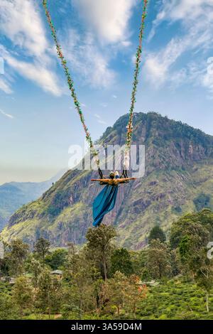 Eine Frau schwingt hoch über den üppigen Hügeln von Ella, Sri Lanka, mit Little Adam's Peak im Hintergrund und genießt ein aufregendes und landschaftlich reizvolles Abenteuer in n Stockfoto