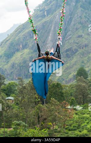 Eine Frau schwingt hoch über den üppigen Hügeln von Ella, Sri Lanka, mit Little Adam's Peak im Hintergrund und genießt ein aufregendes und landschaftlich reizvolles Abenteuer in n Stockfoto