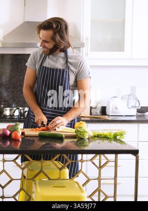 Mann in der Küche, der Gemüse auf der Theke hackt, Schürze trägt und beim Kochen lächelt Stockfoto