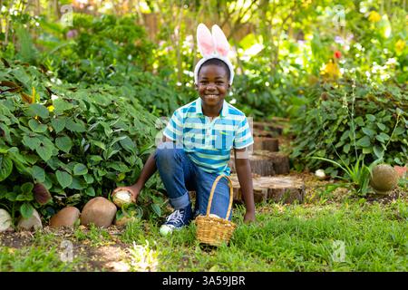 Lächelnder Junge mit Hasenohren, der Ostereier im Garten sammelt Stockfoto