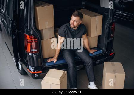 Pause machen, ausruhen, sitzen. Der Lieferer oder Lader befindet sich in der Nähe des Fahrzeugs mit einer Kiste. Stockfoto