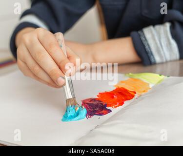 Junge Hand hält einen Pinsel mit blauer Farbe und zeichnet einen Regenbogen auf ein Blatt Papier, ein Junge von 4-5 Jahren zeichnet mit Farben zu Hause. Hochwertige Fotos Stockfoto