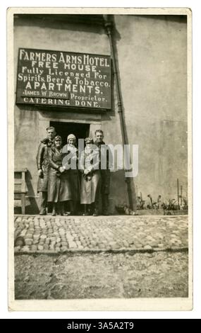 Ursprüngliche Postkarte aus der Zeit der 1920er/frühen 1930er Jahre von männlichen und weiblichen frühen Bikern, auf einer Fahrt, mit langen Ledermänteln, Schutzbrillen, Handschuhen, Kappen. Moderne Frauen! In Großbritannien der 1920er Jahre erlangten Frauen größere Unabhängigkeit, einschließlich des Wahlrechts und des Zugangs zu Berufen, und sie nahmen neue Freiheiten in Mode, Verhalten und sozialem Leben ein, wie sie bei diesen ausgemergelten Frauen zu sehen sind, von denen eine auch stolz raucht. Die Gruppe der Radfahrer sieht aus, als wären sie auf dem Land unterwegs und stehen vor dem Farmers Arms Hotel (James W. Brown ist der Besitzer) irgendwo in Großbritannien Stockfoto