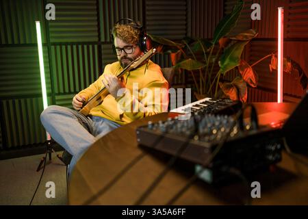 Ein fokussierter männlicher Musiker stimmt eine Gitarre vor dem Hintergrund einer digitalen Klangbearbeitung in einer zeitgenössischen Aufnahmestudioumgebung ab. Stockfoto