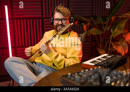 Ein fokussierter männlicher Musiker stimmt eine Gitarre vor dem Hintergrund einer digitalen Klangbearbeitung in einer zeitgenössischen Aufnahmestudioumgebung ab. Stockfoto