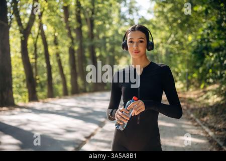 Eine Pause machen. In Kopfhörern und mit Wasser stehen. Die junge sportliche, dünne Frau trägt draußen schwarze Fitnesskleidung. Stockfoto