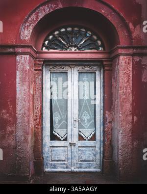 Eine alte weiße Holztür mit Spitzenvorhängen, eingerahmt von einem rot strukturierten Bogengang in einer historischen mediterranen Stadt. Stockfoto