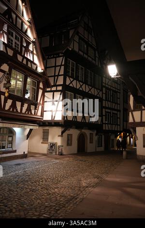 Traditionelle Fachwerkhäuser säumen eine Kopfsteinpflasterallee in Straßburg Petite France, die nachts von Straßenlaternen sanft beleuchtet werden. Stockfoto