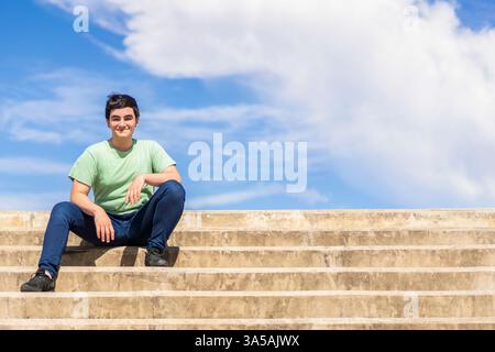 Freundlicher junger Mann, der auf Stufen sitzt und vor einem hellblauen Himmel lächelt Stockfoto