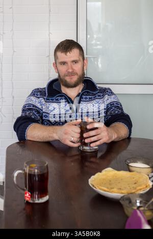 Ein Weißer mittleren Alters mit Bart sitzt an einem Tisch und trinkt Tee aus einer großen Tasse. Das Familienoberhaupt. Stockfoto