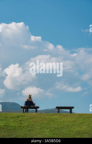 Weibliche Touristen sitzen an einer Holzbank auf einem üppigen grünen Hügel und großen flauschigen Wolken am blauen Himmel an sonnigen Tagen Stockfoto