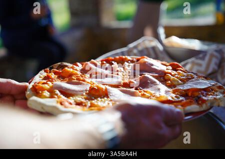 Nahaufnahme einer frisch gebackenen Schinkenpizza, die draußen serviert wird. Die Pizza mit geschmolzenem Käse und dünn geschnittenem Schinken auf knuspriger Kruste ist fertig Stockfoto