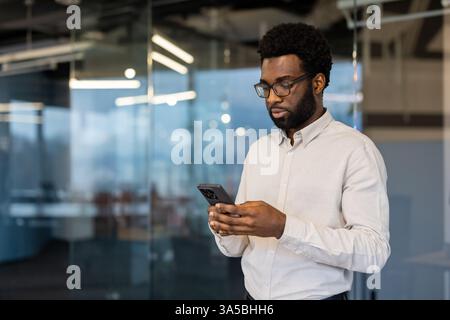 Junger Geschäftsmann im Büro mit Handy. Konzentrieren Sie sich auf einen Mann in einer Brille, der ein Smartphone hält und in modernen Büroräumen online kommuniziert. Stockfoto